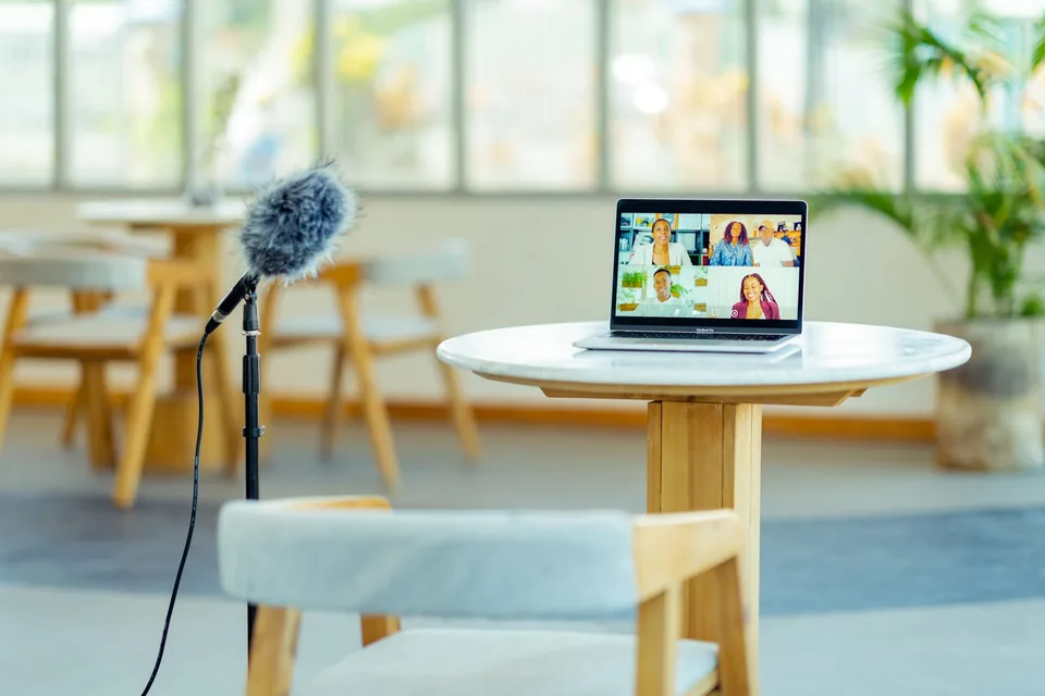 A laptop on a café table showing a video call with five participants, next to a microphone.